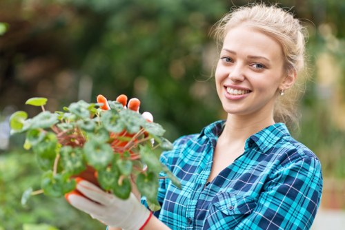 A young woman with blonde hair tied back, wearing a blue plaid shirt and gardening gloves, standing in a well-maintained garden with a lush green lawn, bordered by a row of bushes and small trees in the background. She is holding a potted plant with green leaves and red-tinged foliage, ready to plant or tend it, indicating outdoor gardening activity typical of a residential garden in Dalston. The garden features a combination of natural grass, soil, and some paved or decking areas, with diffuse daylight suggesting a mild, partly cloudy day suitable for gardening services offered by Landscapers Dalston. The environment appears clean, organized, and inviting, reflecting the outdoor maintenance and landscaping expertise provided locally in Dalston and surrounding areas.