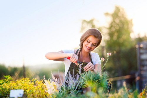 A woman smiling while tending to a garden with a variety of plants, including flowering herbs and green foliage, in a well-maintained outdoor space. She appears to be watering or planting, with a garden bed bordered by a mix of flowering perennials and lush green shrubbery. The background features a softly blurred garden scene with trees and a partly cloudy sky, suggesting a bright, pleasant weather day. The garden has a neatly arranged layout with defined planting areas, and the surface includes soil beds and possibly some decorative paving, indicating professional landscaping work from a company like Landscapers Dalston. The natural tones of green and soft floral colours highlight the healthy garden environment suitable for outdoor gardening and maintenance services in Dalston or nearby London neighborhoods.