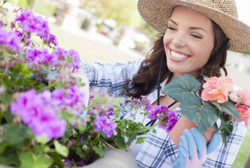A woman wearing a wide-brimmed straw hat and light blue gardening gloves is smiling as she tends to a variety of colorful flowering plants in a well-maintained garden. The garden features lush green grass, flower beds filled with vibrant pink and purple blooms, and a backdrop of trees and a clear sky, suggesting a bright, sunny day. The scene is set in a landscaped outdoor space, possibly part of a front or back garden in Dalston, with visible natural textures such as soil, plant foliage, and decorative planting containers. This detailed visual highlights outdoor gardening activities and can support services offered by landscaping and gardening professionals like Landscapers Dalston.