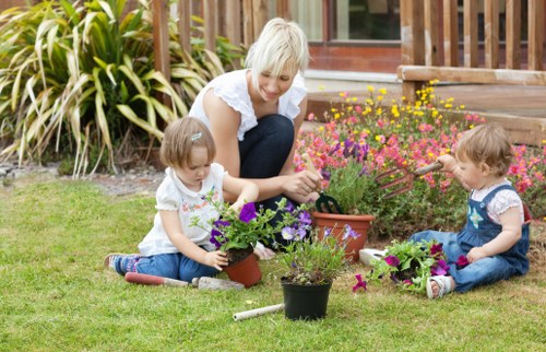 In a well-maintained garden in Dalston, a woman with short blonde hair is engaging in planting activities alongside two young children. The garden features a lush, green lawn with dense grass and a variety of flowering plants, including vibrant pink, purple, and yellow blossoms arranged in flower beds near a wooden deck. The children, wearing casual clothes, are sitting on the grass and holding small pots filled with colorful flowers, actively participating in gardening. The background includes a wooden fence and a small pathway leading to a deck area, with tall ornamental grasses bordering part of the garden. The scene is lit by natural daylight, indicating a clear and bright day, creating a cheerful outdoor environment. This setting highlights the importance of garden maintenance and outdoor family activities, relevant to the gardening services offered by Landscapers Dalston, who can assist with lawn care, planting, and overall outdoor space enhancement in Dalston and nearby areas.