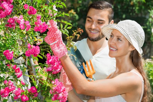 A smiling man and woman are working together in a garden, surrounded by lush greenery and vibrant pink flowering bushes. The woman, wearing a white hat with black polka dots and pink gardening gloves, is holding a small pruning shears and appears to be trimming the pink flowers. The man stands behind her, dressed in casual outdoor clothing, observing her work with a pleased expression. Their garden features a dense hedge or shrubbery in the background, along with a variety of green foliage. The scene is brightly lit by natural sunlight, indicating clear weather, and the overall environment suggests a well-maintained outdoor space suitable for gardening services in the Dalston area. This image demonstrates typical garden maintenance activities such as pruning and plant care, aligning with landscaping and lawn care services provided by Landscapers Dalston.