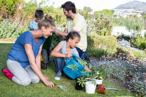 A family gardening in a well-maintained backyard with a neatly trimmed lawn, surrounded by flowering plants and shrubs, and bordered by a low hedge. In the foreground, a woman is kneeling on the grass, planting or tending to a small shrub, with gardening tools and potted plants nearby. A young girl sits on the grass, holding a watering can, while a man and a boy stand in the background, engaging with each other amidst the garden's lush greenery. The outdoor space appears to be part of a residential property in Dalston, with a view of distant hills and houses under an overcast sky, suggesting mild weather. The garden features a mix of flower beds with natural multicolour blooms, as well as patches of vibrant green grass with dense, healthy turf. The scene captures a moment of family engagement in outdoor landscaping or gardening activities, supported by professional services like those offered by Landscapers Dalston, to help maintain attractive and functional garden spaces.