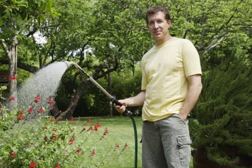 A man stands in a lush, well-maintained garden surrounded by vibrant greenery, including a neatly trimmed hedge, flowering plants, and a lush lawn. He is wearing a light yellow t-shirt and grey cargo shorts, smiling as he holds a garden hose, watering the plants. In the background, mature trees provide shade, and the garden features a mix of flower beds with small red and pink blooms, along with dense shrubbery. The outdoor space appears to be part of a private garden in Dalston, with natural sunlight highlighting the varied textures of the grass, soil, and plant foliage. The scene exudes a peaceful, well-kept environment typical of professional gardening and landscaping services, such as those offered by Landscapers Dalston, aimed at maintaining healthy and attractive outdoor spaces.