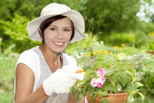 A woman with short brown hair, wearing a wide-brimmed white hat, white gardening gloves, and a light-colored short-sleeved shirt, is smiling while tending to a potted flowering plant in a lush outdoor garden. The garden features a well-maintained lawn with dense green grass, bordered by flower beds containing various colorful blooms and shrubs. In the background, there are taller trees and bushes providing a natural, layered landscape. The weather appears bright and sunny, with soft natural light illuminating the scene. The woman is carefully handling the plant, which has pink and white flowers, indicating she is engaged in gardening or plant care activities typical to outdoor landscape maintenance. This setting reflects a typical well-kept garden space suitable for professional gardening services offered by landscapers Dalston, emphasizing vibrant plant life, tidy surfaces, and a lush natural environment within a residential or public outdoor area in London postcode area, N16.