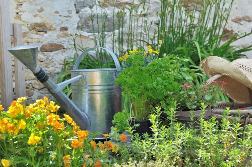 In a well-maintained garden, there is a variety of plants and flowers growing close to a weathered stone wall. The foreground features vibrant yellow and orange flowers, providing bright colour accents against the lush green foliage. Behind the flowers, there are several potted plants, including leafy greens and flowering varieties, arranged on the ground. A classic metal watering can and a small, aged garden hat rest among the plants, indicating ongoing garden care. Tall, slender plants with narrow leaves extend upward, possibly flowering perennials or herbs. The surface beneath the plants appears to be loose soil or mulch, supporting the healthy growth of the garden. The environment is outdoors with natural daylight, suggesting a peaceful setting suitable for gardening and outdoor maintenance. This scene exemplifies typical garden features relevant to gardening services in Dalston, where careful plant arrangement and outdoor tools contribute to a vibrant, cultivated yard. The overall composition reflects a tidy, thriving garden space designed for ongoing horticultural care and landscaping opportunities by local specialists.