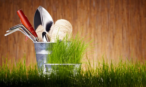 A close-up of gardening tools and supplies placed in a small metal bucket outdoors on a lush green lawn. The bucket contains various garden implements including a red-handled trowel, a black-handled hand fork, a pair of pruning shears, and a spool of natural twine. The surrounding grass is vibrant and freshly mowed, with individual blades clearly visible in the foreground. In the background, there is a wooden fence with a warm natural finish, indicating a typical garden setting in Dalston, East London. The overall scene suggests preparation for gardening or plant maintenance activities, and the natural lighting indicates a bright, possibly sunny day suitable for outdoor gardening work. This image visually supports garden maintenance and landscaping services provided by Landscapers Dalston, emphasizing tools and environment conducive to lawn and garden care within an outdoor residential space.