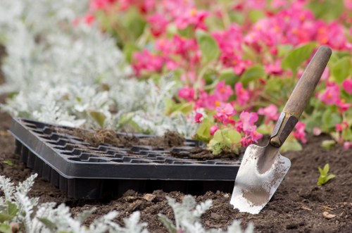A close-up image of a gardening scene taken outdoors on a cloudy day in Dalston, showcasing a freshly cultivated flower bed with dark, moist soil. In the foreground, a small gardening trowel with a wooden handle and metal blade is partially inserted into the earth. Behind the trowel, a black seed tray sits on the soil, containing small seed-starting pots with soil and some young seedlings. Surrounding the flower bed are lush, low-growing plants with silvery-green foliage, creating a natural border. In the background, vibrant pink and white flowering plants are visible, adding colour to the landscaped garden. The overall scene captures a moment of planting or soil preparation, illustrating professional gardening services that focus on floral and bed planting in outdoor spaces within the local area, supported by Landscapers Dalston's expertise.