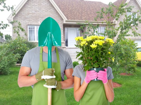 In a residential garden setting, a person wearing an open-front green gardening apron and light-colored gloves holds a shovel with a teal plastic cover over the blade, positioned vertically in front of their body. Next to them, another person wearing a matching green apron and pink gardening gloves holds a vibrant potted plant with bright yellow flowers and lush green foliage, obscuring their face. The garden features a well-maintained lawn with dense, green grass, bordered by a variety of shrubs and small trees. In the background, a light-colored brick house with white-framed windows and a pitched roof is visible, framed by a few trees and bushes. The scene appears to be outdoors on a clear day, with natural daylight illuminating the garden and the individuals. The setting suggests activities related to gardening or landscape enhancement, supported by the presence of gardening tools and plants, consistent with services provided by Landscapers Dalston in the local area.